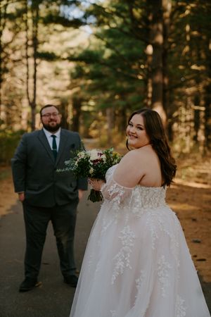 Bride in off-shoulder lace gown poses with groom in forest setting.
