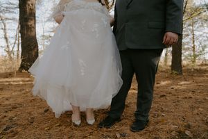Close-up of wedding attire showing a flowing white dress and dark suit against a forest backdrop.