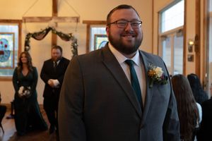 A groom in a gray suit and glasses stands smiling during a wedding ceremony.