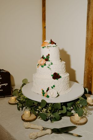 Three-tiered white wedding cake decorated with burgundy and peach flowers on a greenery-adorned stand.
