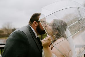 Newlyweds sharing a romantic kiss under a clear umbrella on their rainy wedding day.