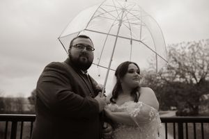 Black and white portrait of newlyweds posing with clear umbrella on outdoor deck.