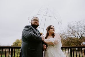 Wedding couple standing together on a balcony holding a clear umbrella against cloudy sky.