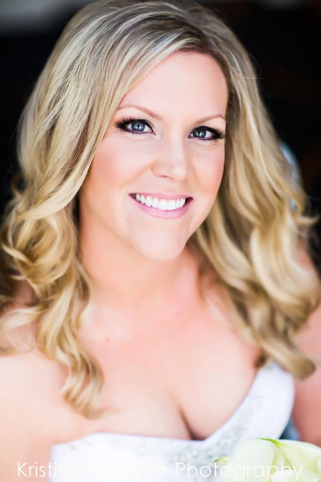 Portrait of a smiling blonde woman with wavy hair wearing a white strapless top against a dark background.