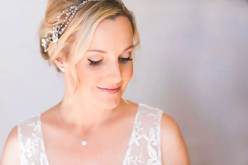 Bride in white lace wedding dress with elegant updo hairstyle poses against soft neutral background.