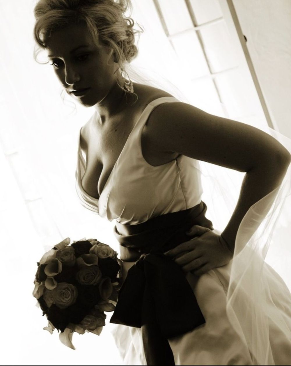 Artistic sepia portrait of a bride in white dress with dark sash holding bouquet by window light.