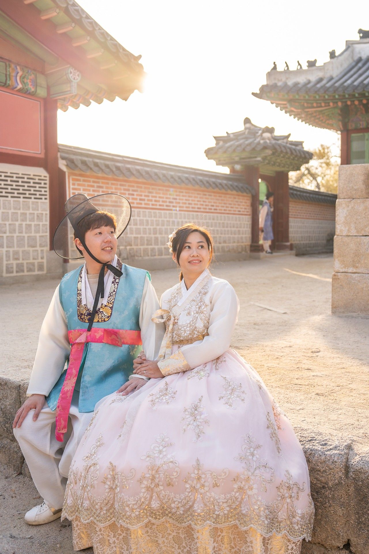 A couple in traditional Korean hanbok attire sits together on stone steps during a sunny outdoor photoshoot.