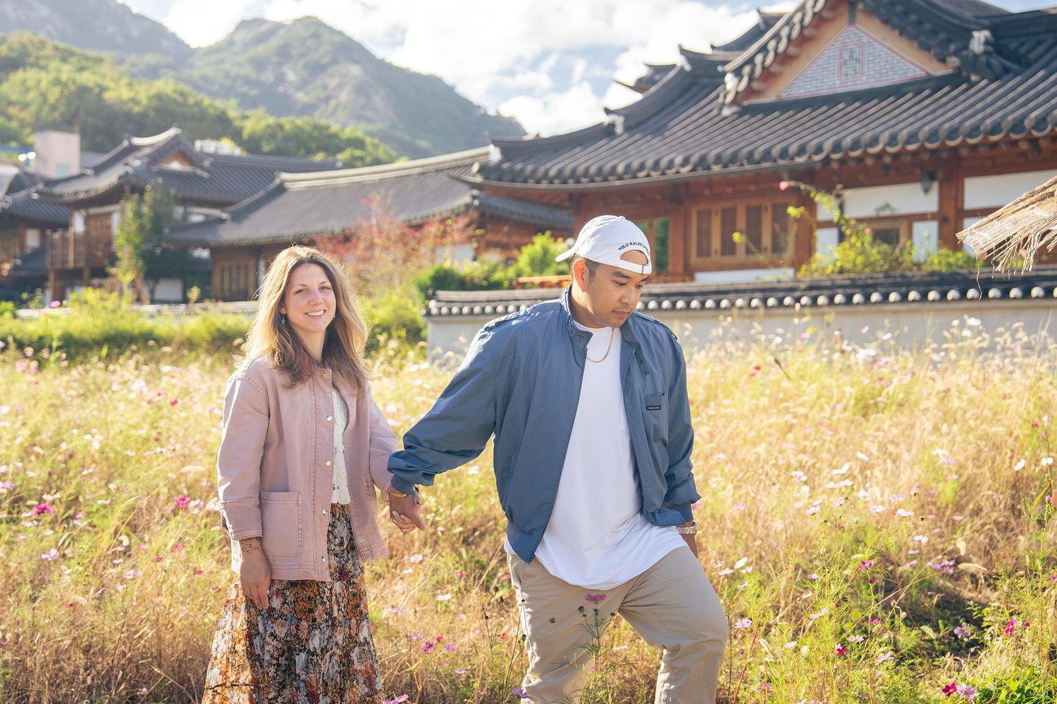 Two people walking through a scenic autumn field with traditional Korean hanok buildings and mountains in the background.
