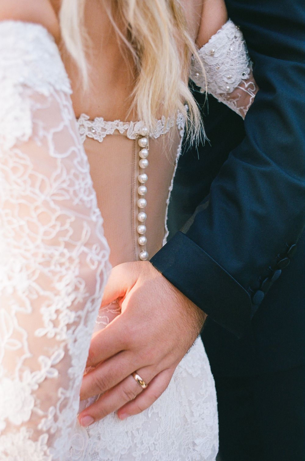 Close-up details of a white lace wedding dress with pearl buttons and hands showing a gold wedding band.