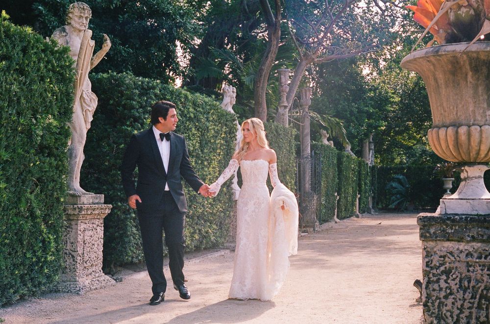 A couple walks hand in hand along a romantic garden path lined with statues and greenery on their wedding day.