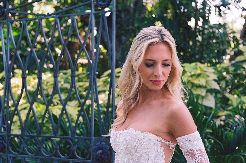 Bride in off-shoulder lace gown poses in garden setting with ornate iron fence and greenery.