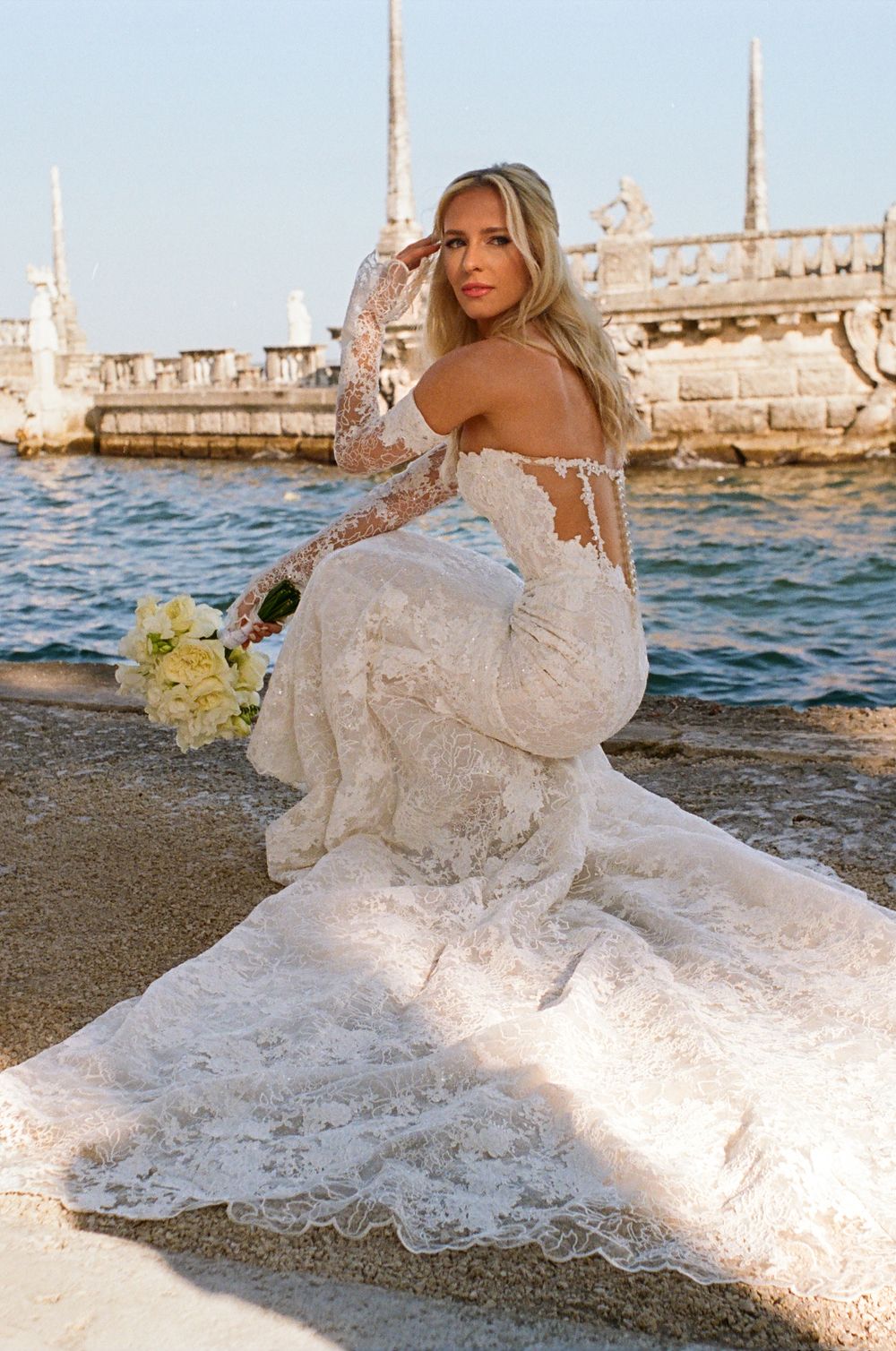 A bride in a lace wedding dress poses by the water with a bouquet near ornate architecture.