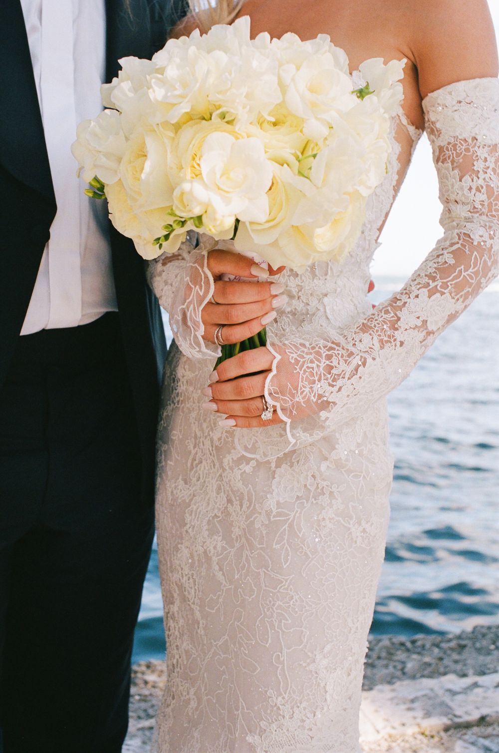 Close-up detail shots of a white lace wedding dress with delicate floral patterns and a cream-colored bouquet by the water.