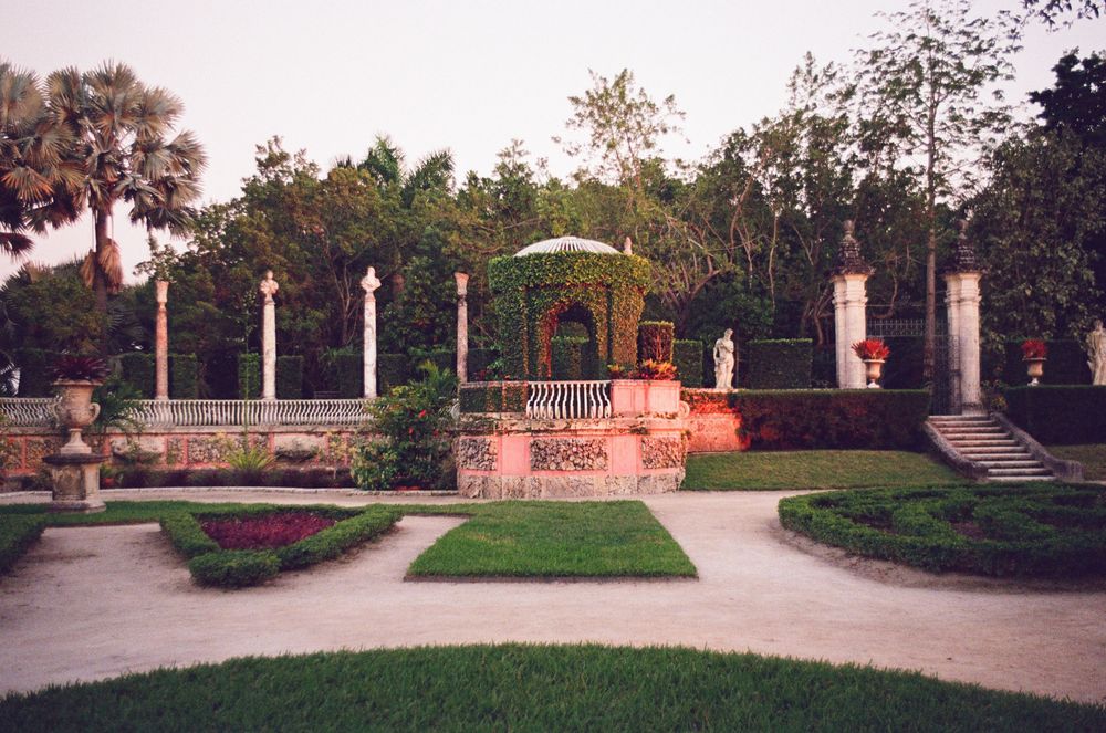 A decorative gazebo with white columns and landscaped gardens in a formal park setting at sunset.