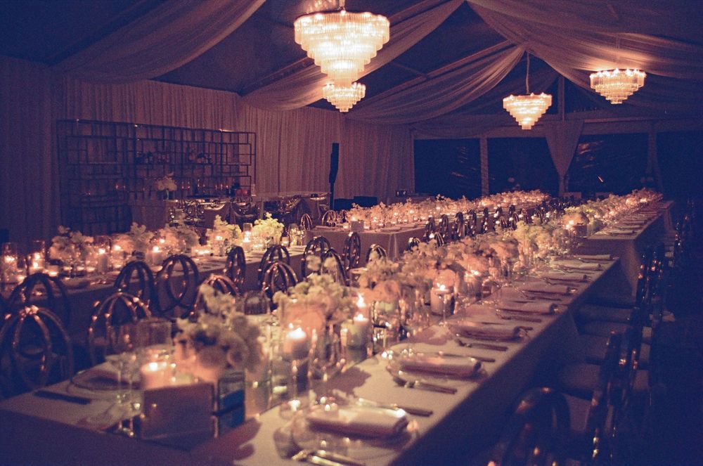 Elegant wedding reception with long dining table, crystal chandeliers, candlelight, and floral centerpieces in a draped tent venue.