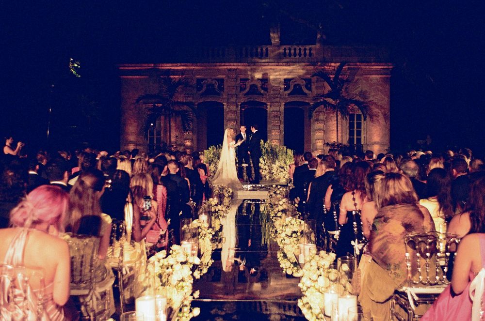 Elegant outdoor wedding ceremony at night with floral aisle, candles, and grand illuminated building backdrop.