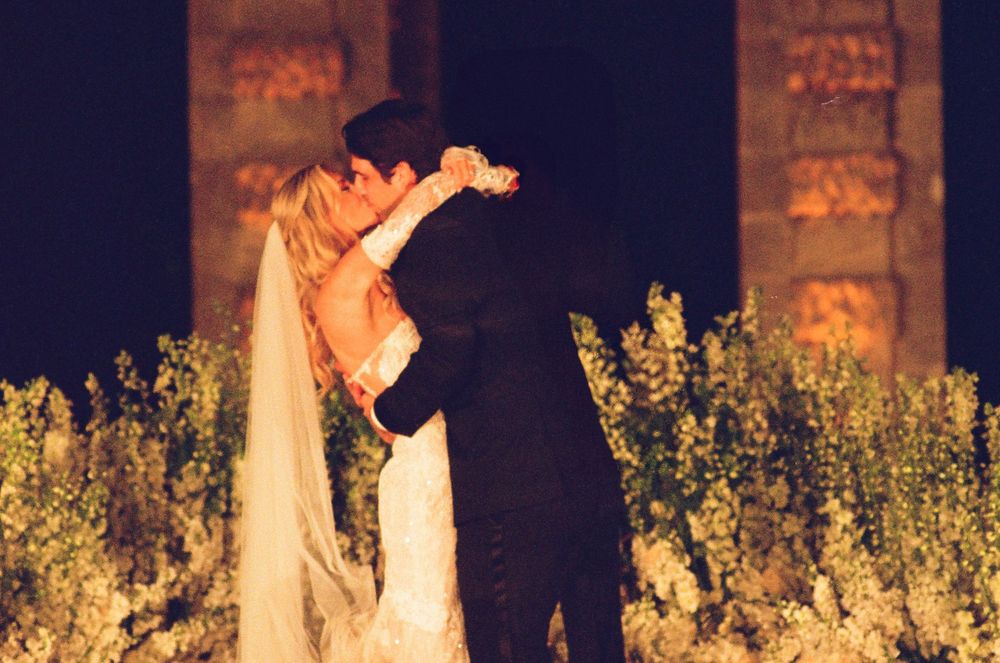 A couple embraces and kisses in a garden at night with illuminated stone walls and yellow flowers in the background.