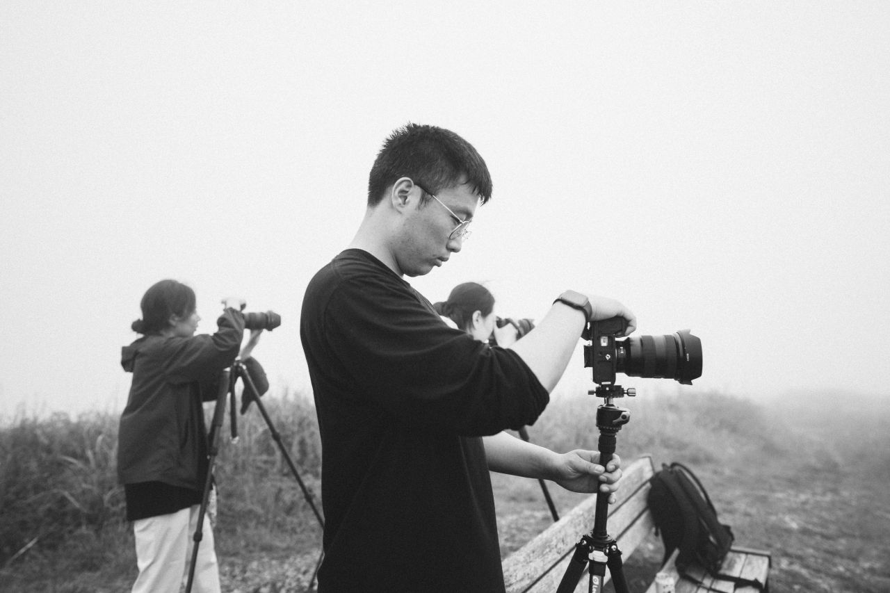A group of photographers stand on a misty mountain ridge with cameras and equipment to capture landscape shots.