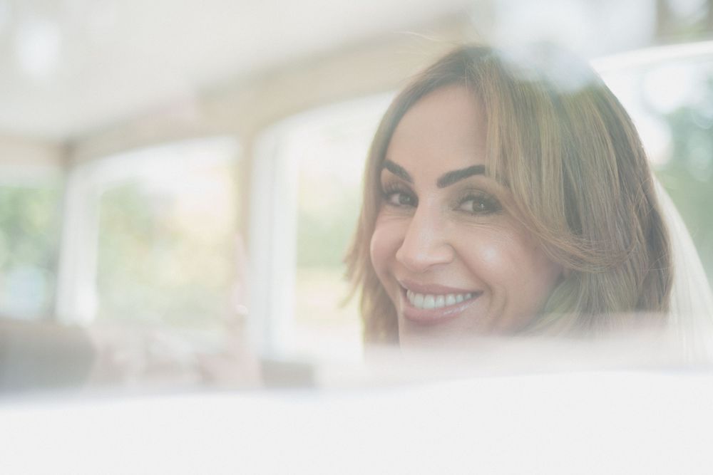 A smiling woman with blonde hair gazes through a window, radiating warmth and confidence in a bright, airy setting.