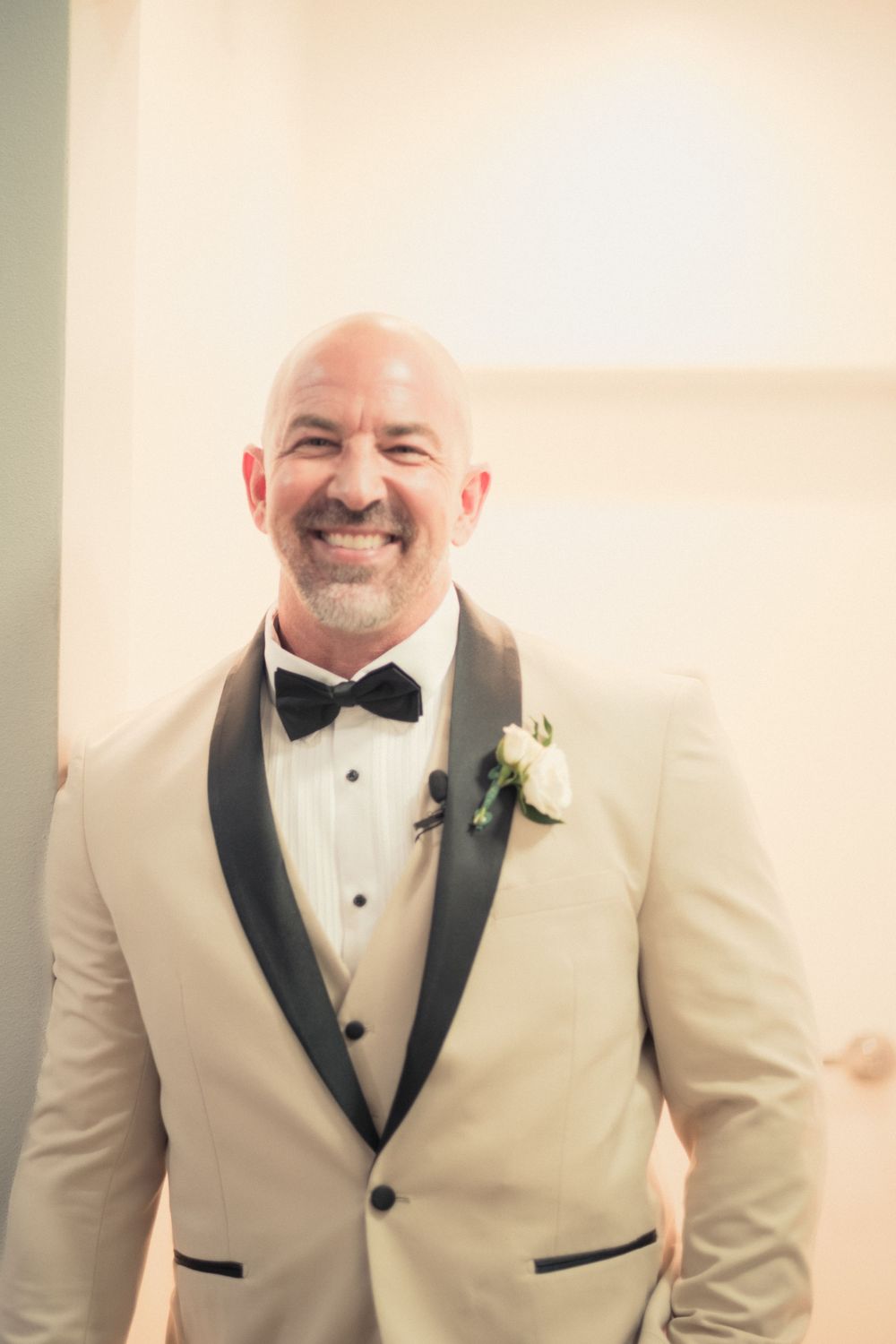 Smiling bald groom in cream tuxedo with black lapels and white boutonniere poses at wedding ceremony.