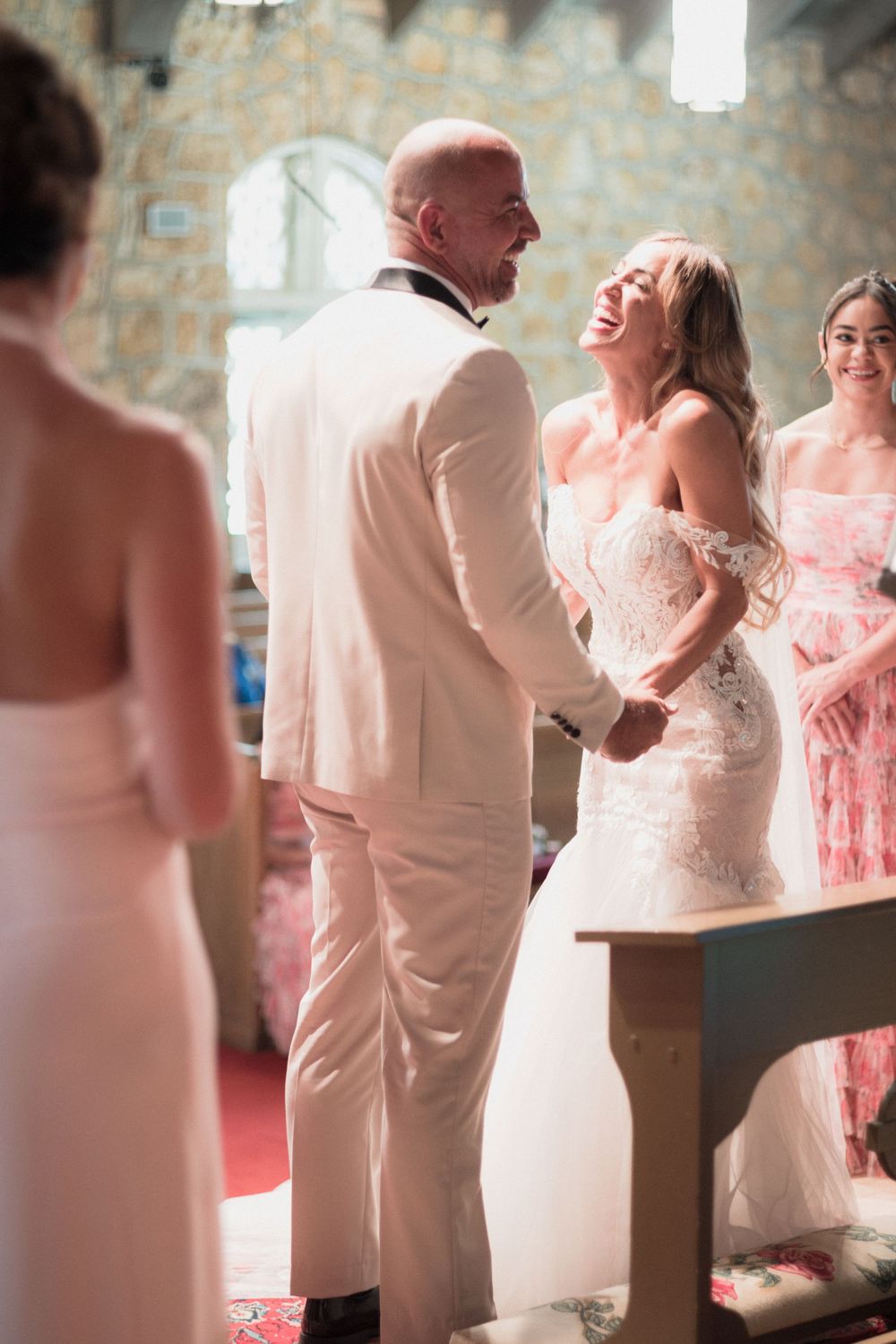 Bride laughing joyfully with groom in white suit during wedding ceremony inside a stone church.
