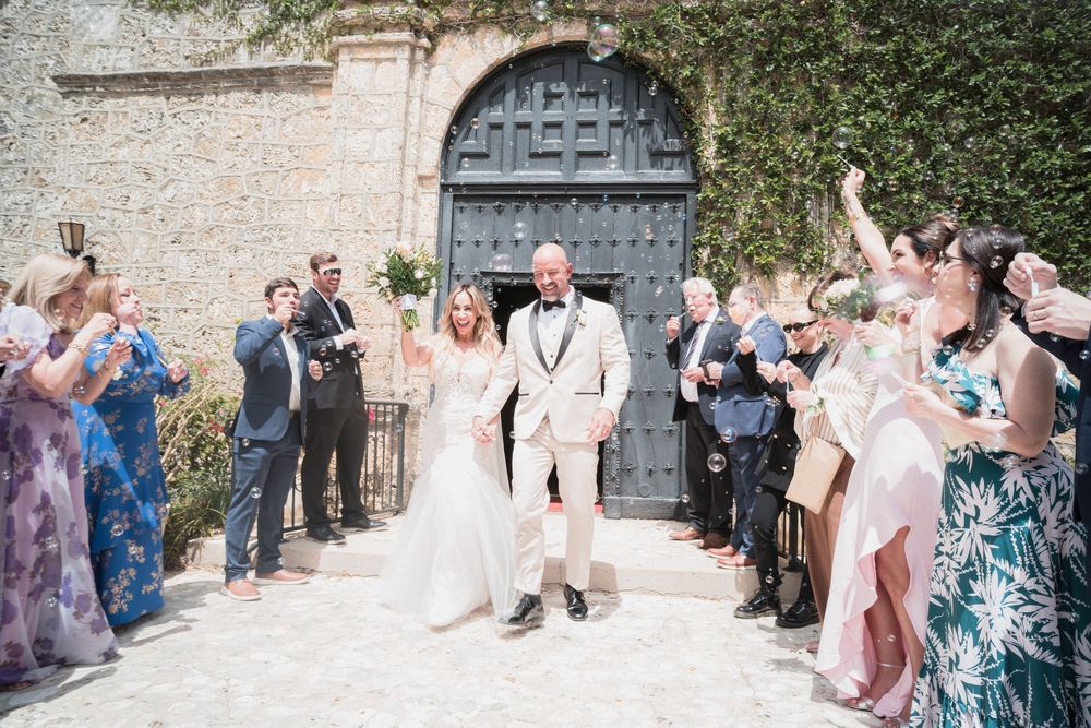 Bride and groom exit church doors as guests toss confetti during sunny outdoor wedding celebration.