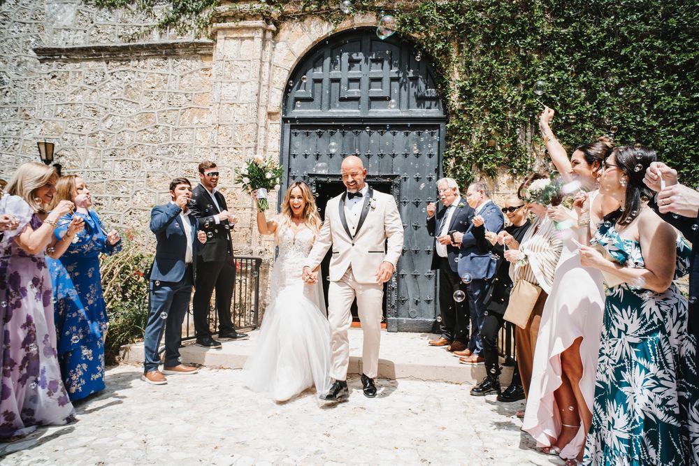 Wedding guests throw confetti at the newlyweds outside a historic stone church with ivy-covered walls.