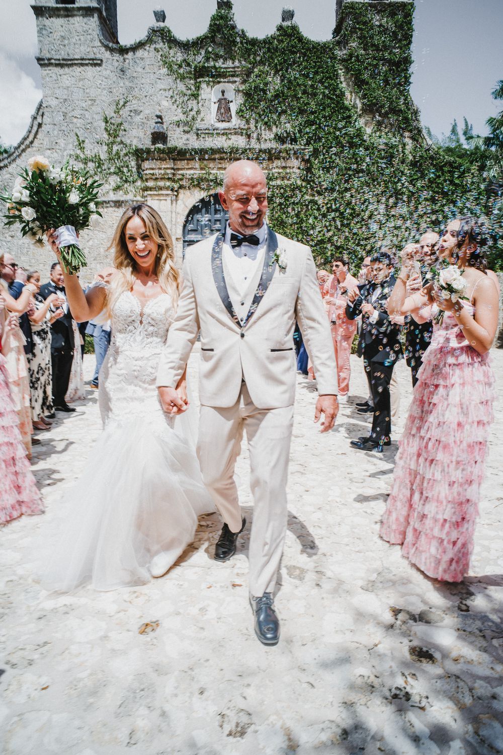 Wedding guests shower newlyweds with confetti as they walk through a stone courtyard in white attire.