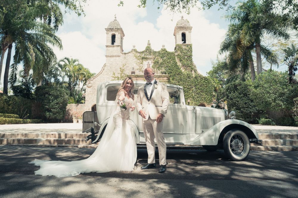 Bride and groom posing with a vintage white car in front of a historic church surrounded by palm trees.