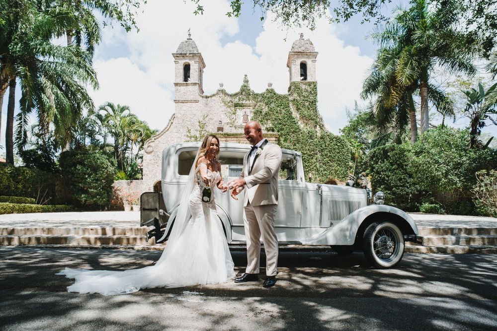A wedding couple poses in front of a white colonial church with palm trees framing the romantic scene.