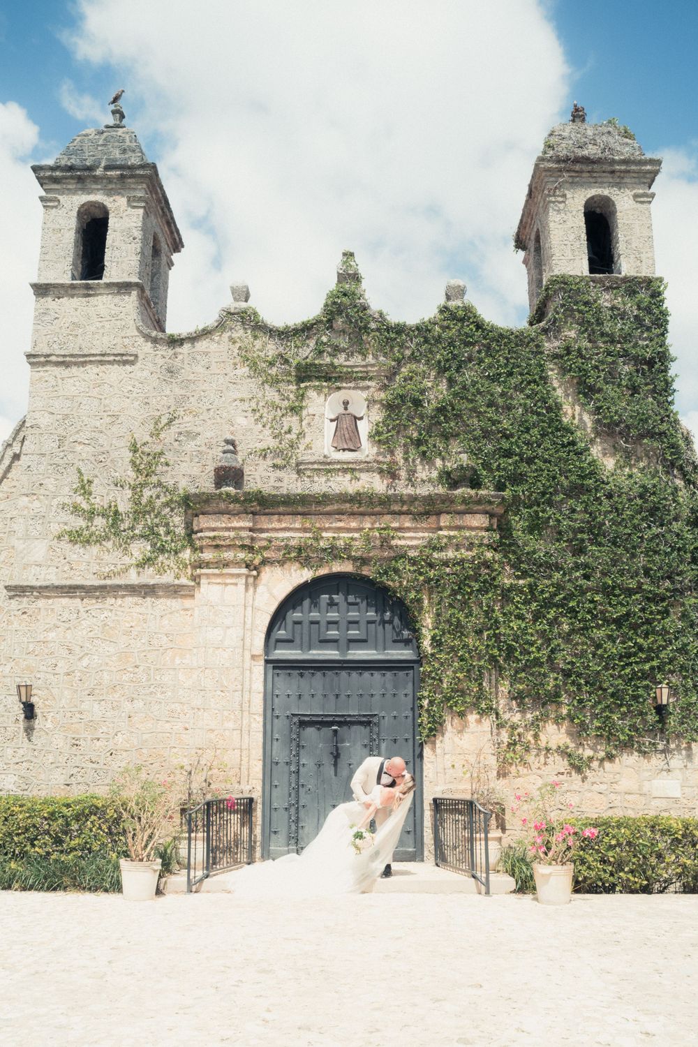 Bride and groom share a romantic dip kiss in front of a historic ivy-covered stone church with twin bell towers.
