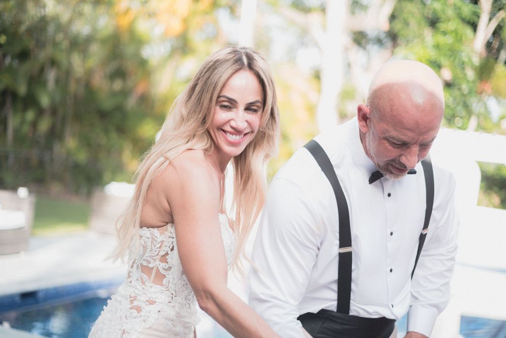 Smiling bride in lace dress stands beside bald groom in white shirt with suspenders at outdoor wedding.