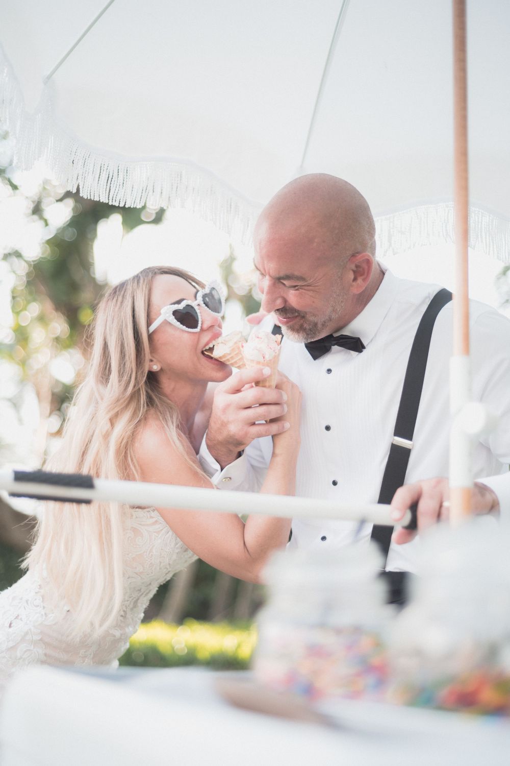 Bride and groom sharing an ice cream cone under a white umbrella at their outdoor wedding reception.