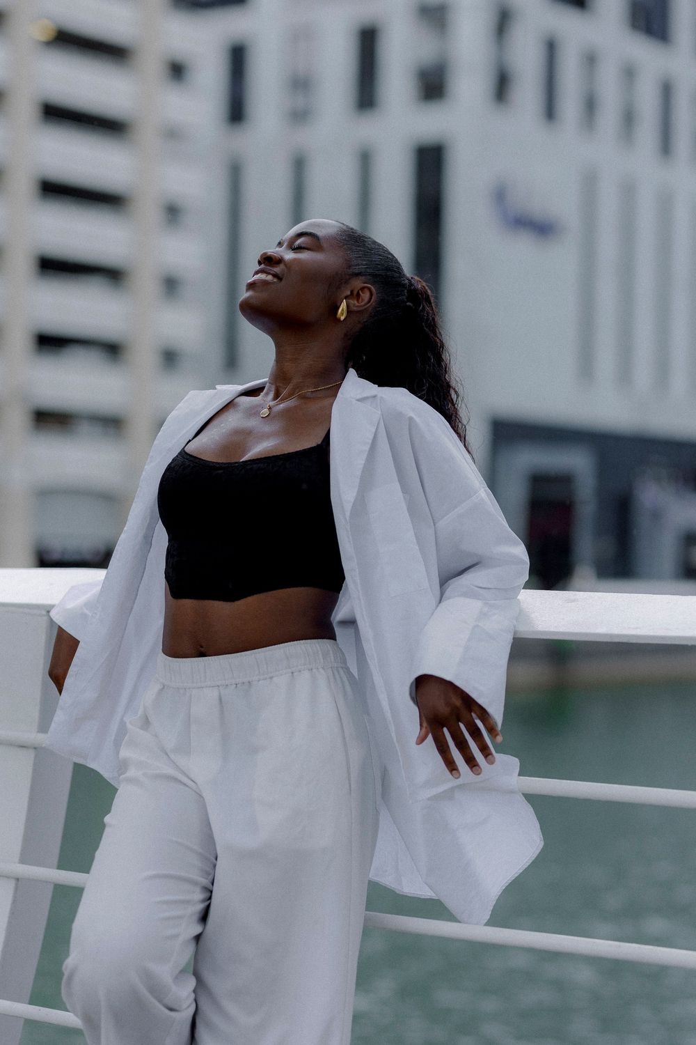 Woman in stylish white blazer and pants with black crop top poses confidently against urban backdrop near water.