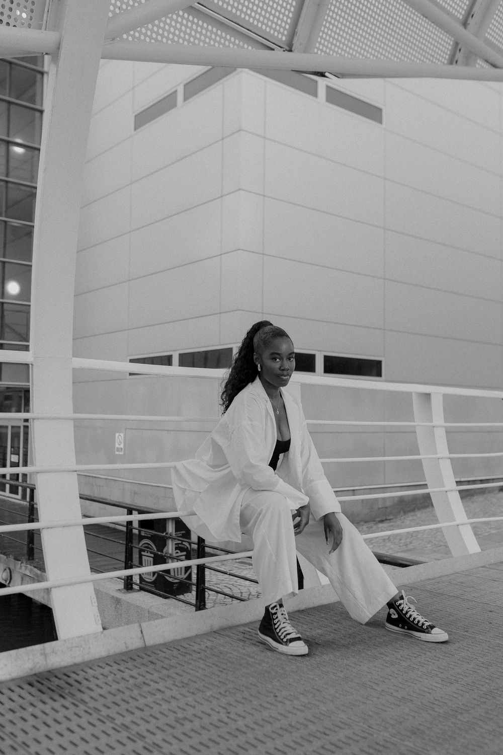 Black and white photo of a person in a white outfit and sneakers sitting on railing in a modern architectural space.