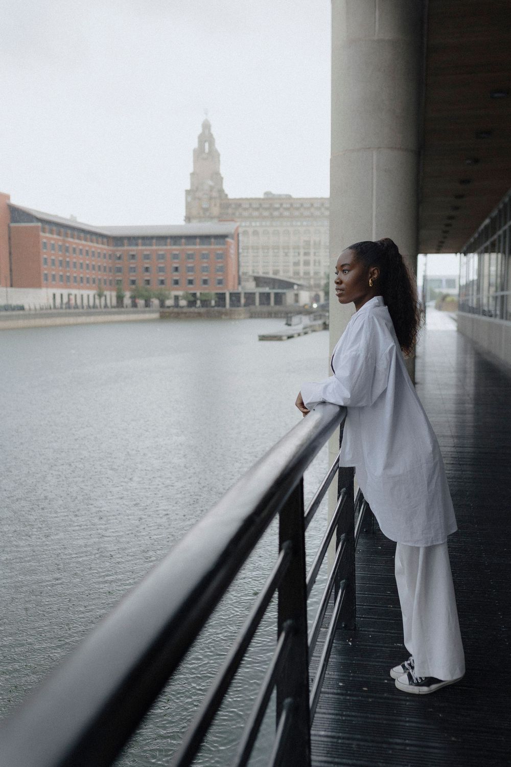 A figure in white clothing leans against a railing overlooking a waterfront cityscape on a cloudy day.