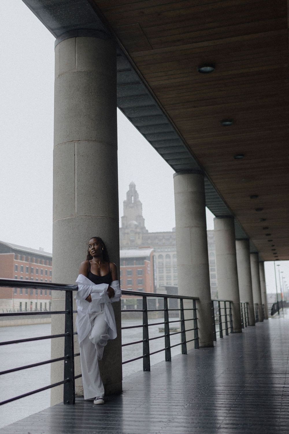 A figure in a white coat stands in a covered walkway with columns and railings overlooking a cityscape.