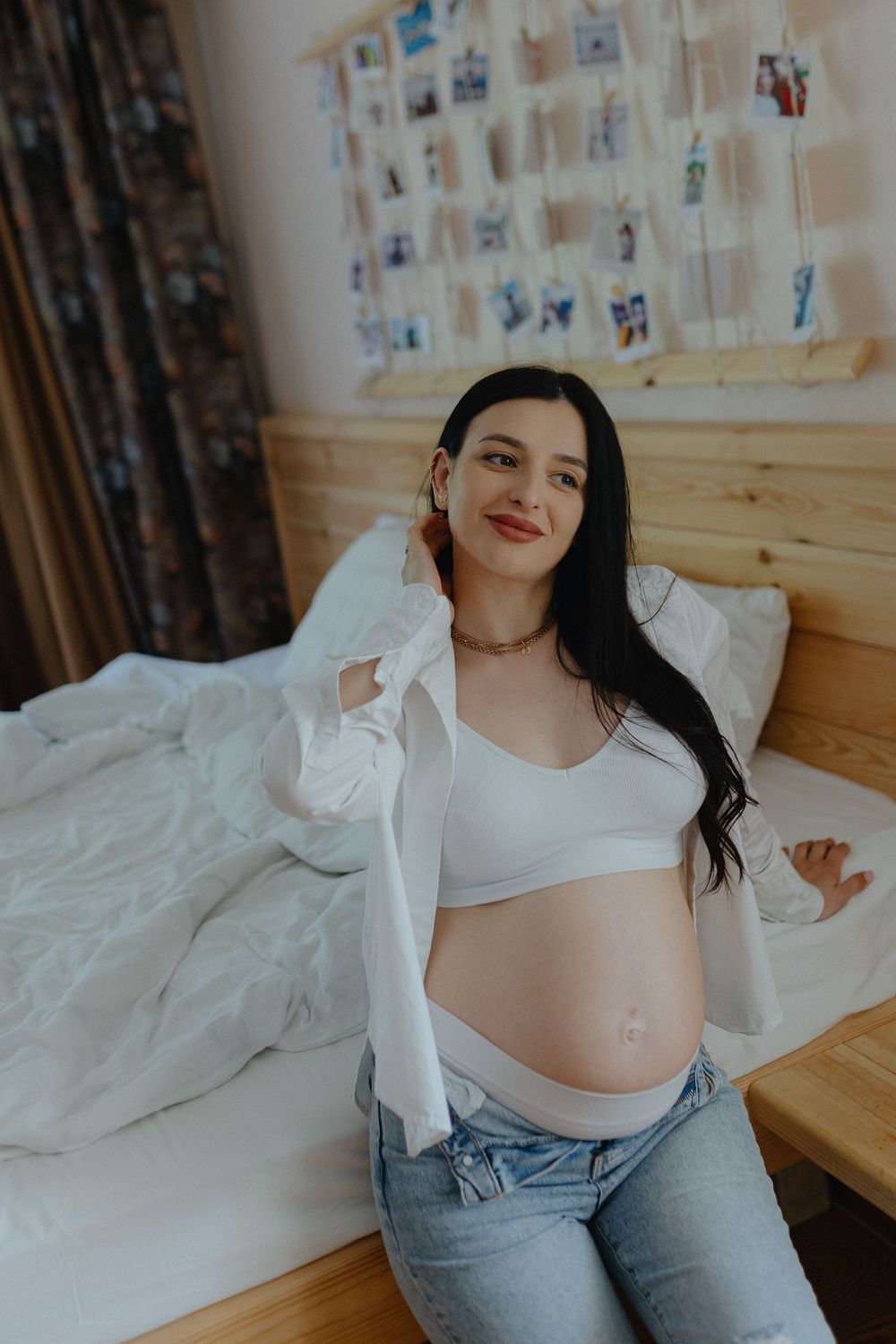 Casual indoor portrait showing someone relaxing on bed wearing white shirt and jeans with photos displayed on wall.
