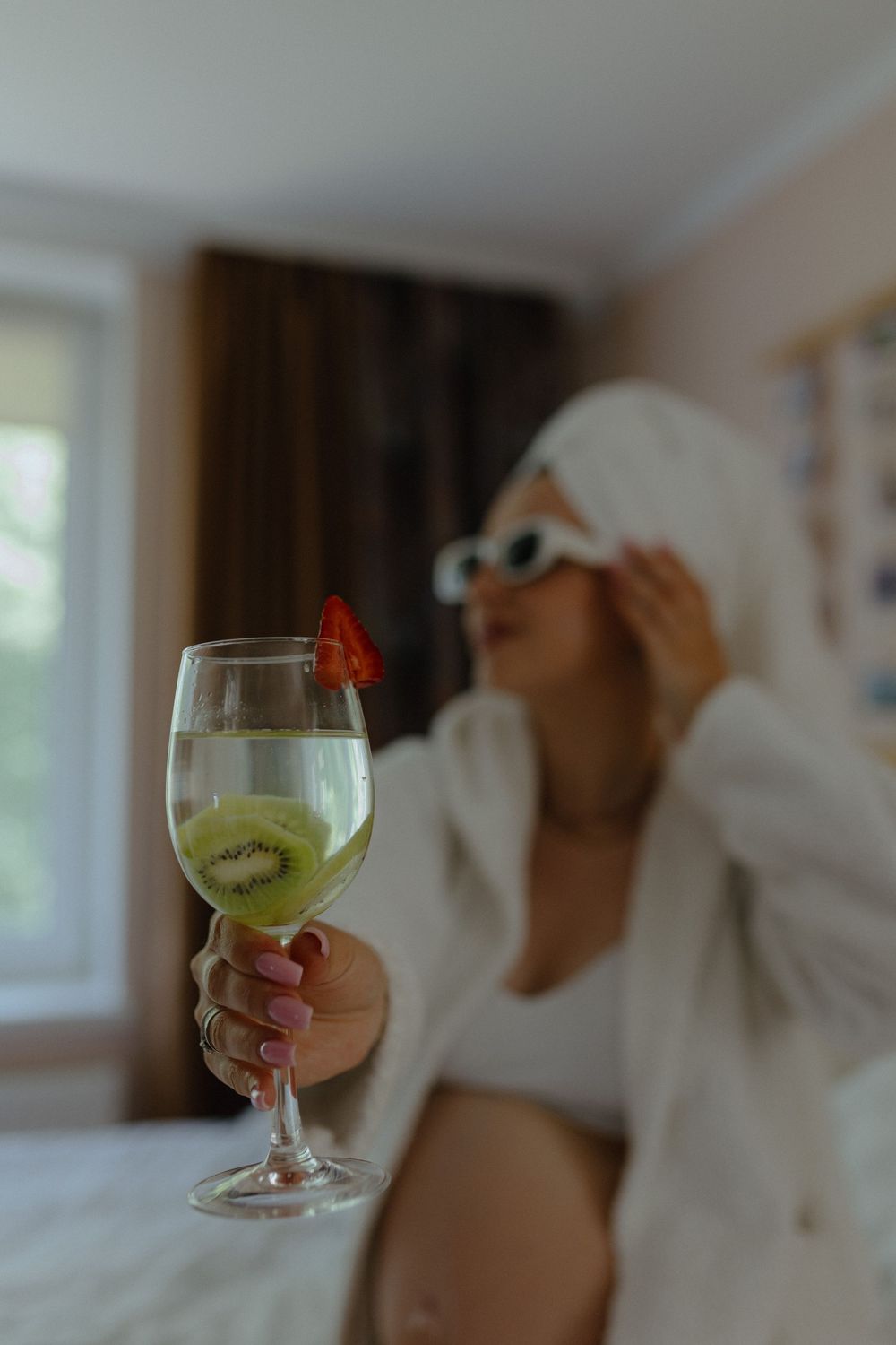 A hand holding a wine glass with a green beverage and sliced kiwi fruit garnish against a blurred background.