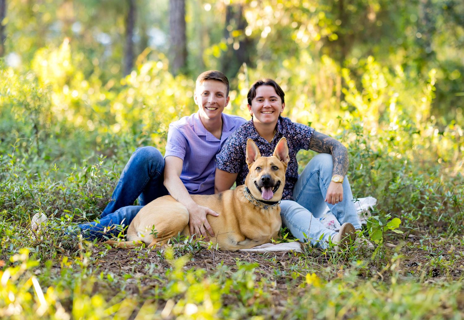A couple and their German Shepherd dog relax together on autumn leaves in a sunlit outdoor setting.