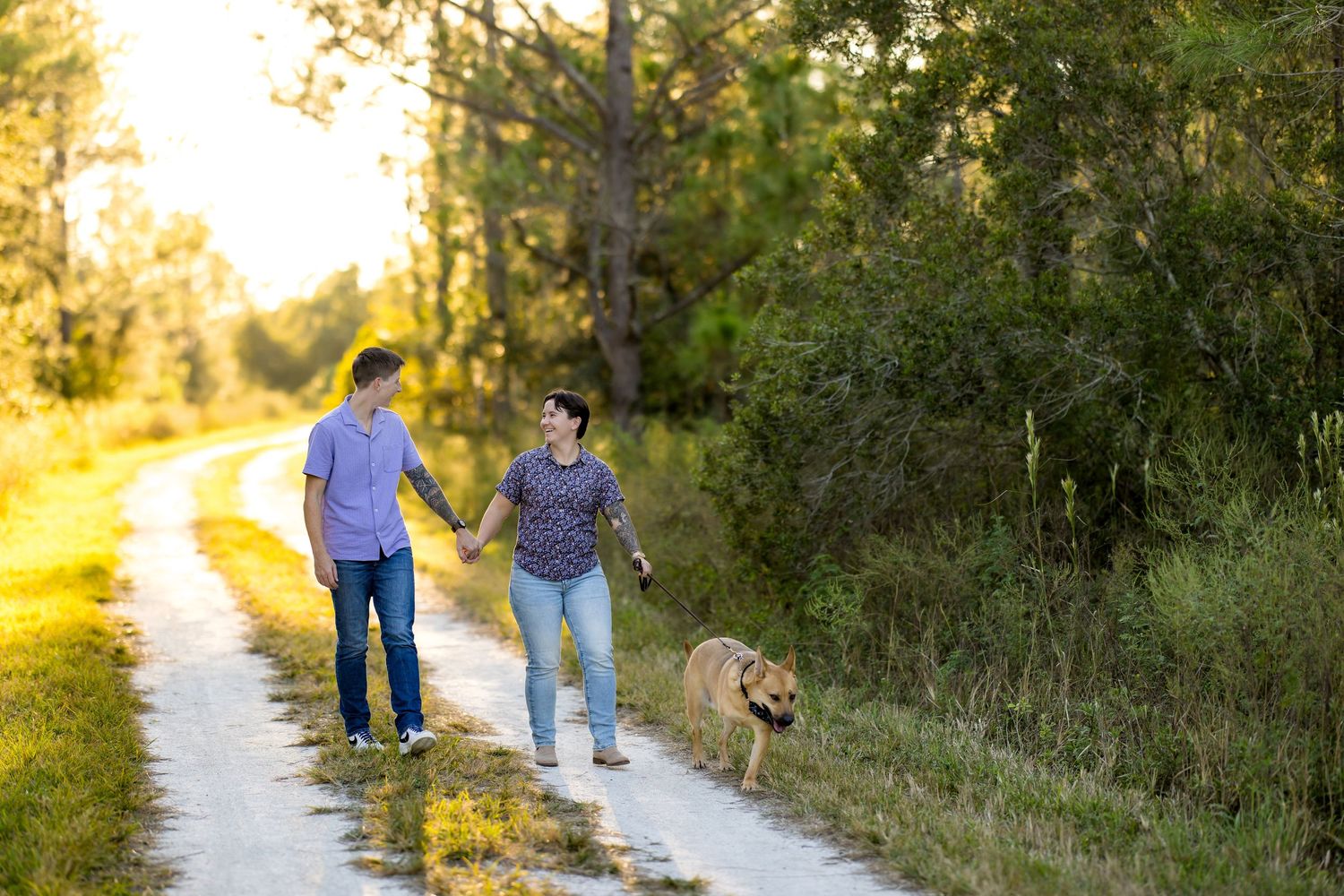 People walk with their golden retriever dog along a sunlit nature trail during golden hour.