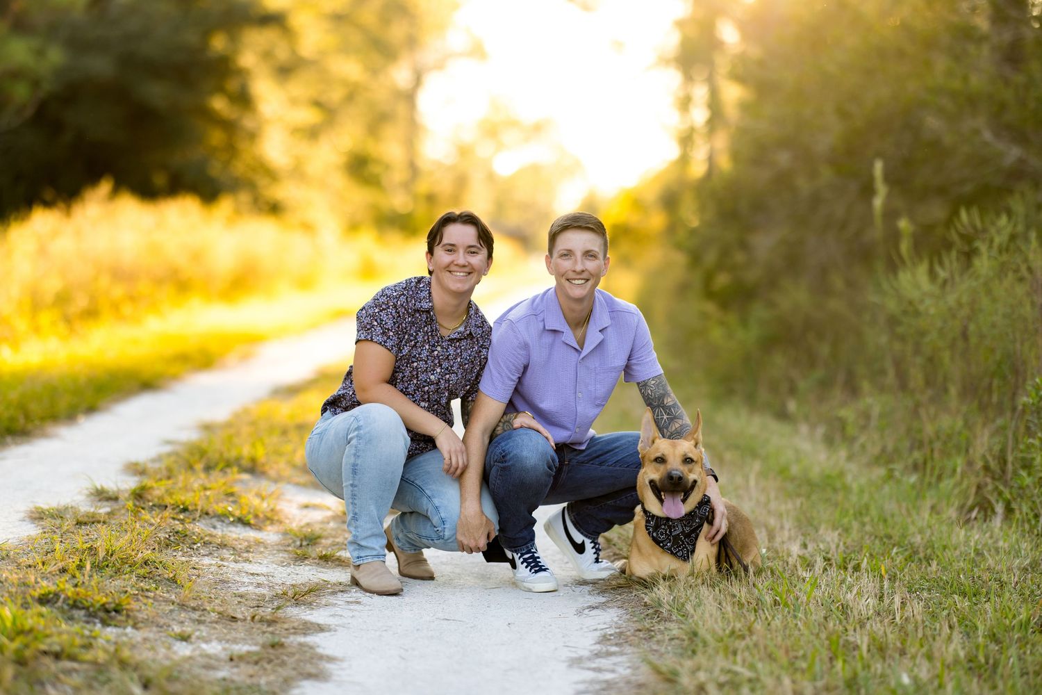 A couple crouches on a sunlit dirt path with their German Shepherd dog during golden hour.