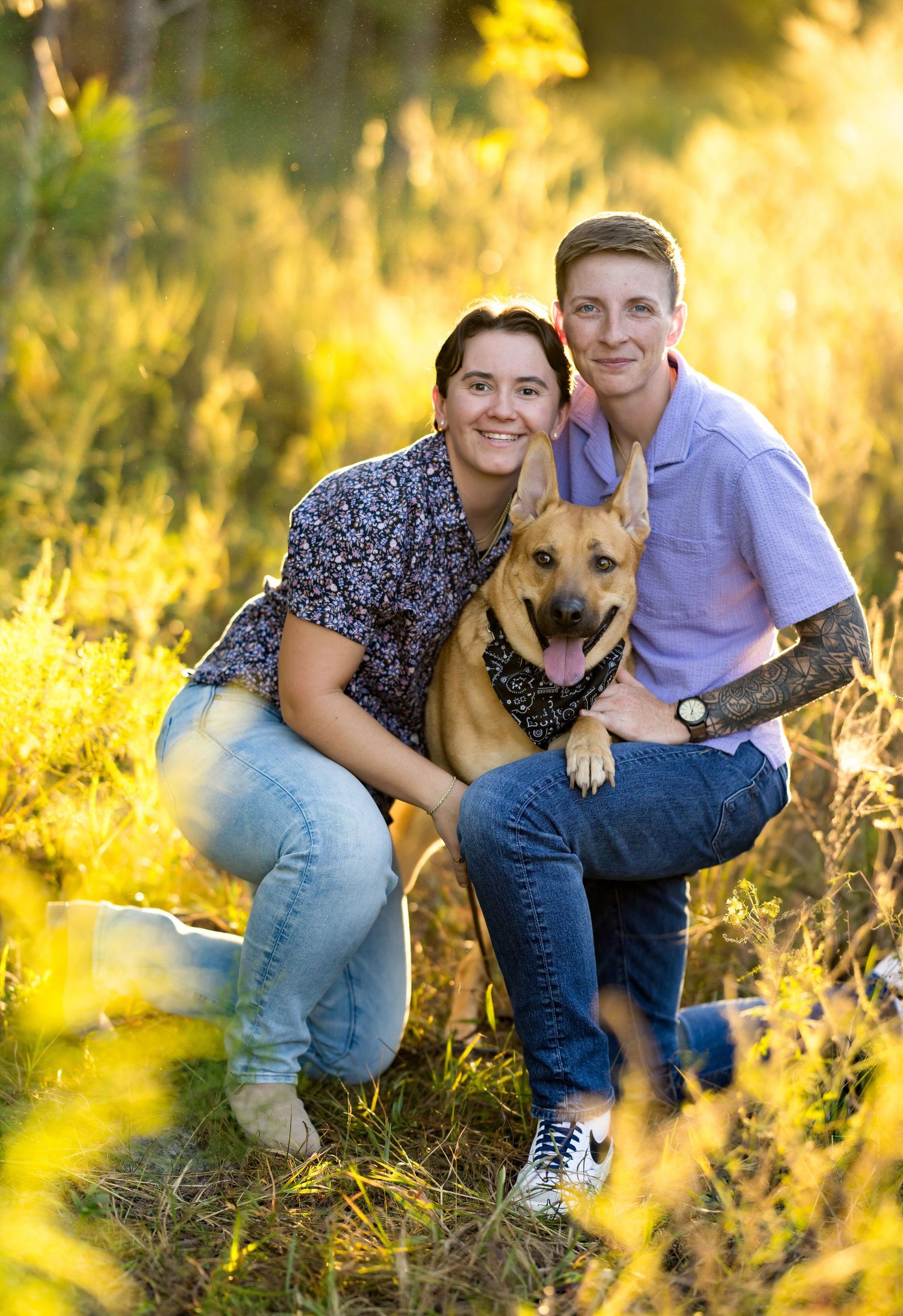 A couple poses with their German Shepherd dog in a sunlit field during golden hour.