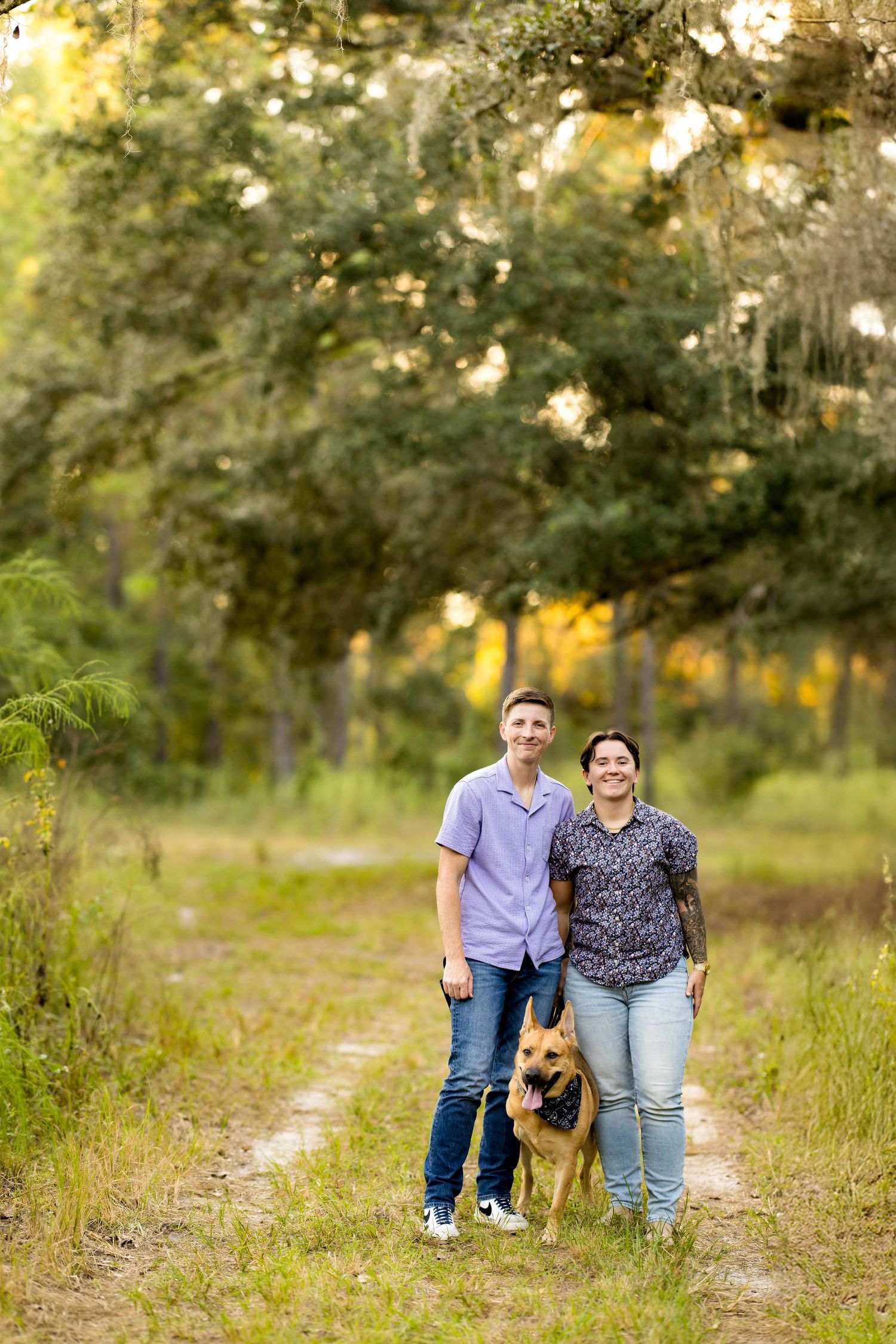 A couple and their German Shepherd dog pose together on a nature trail during a golden sunset photoshoot.