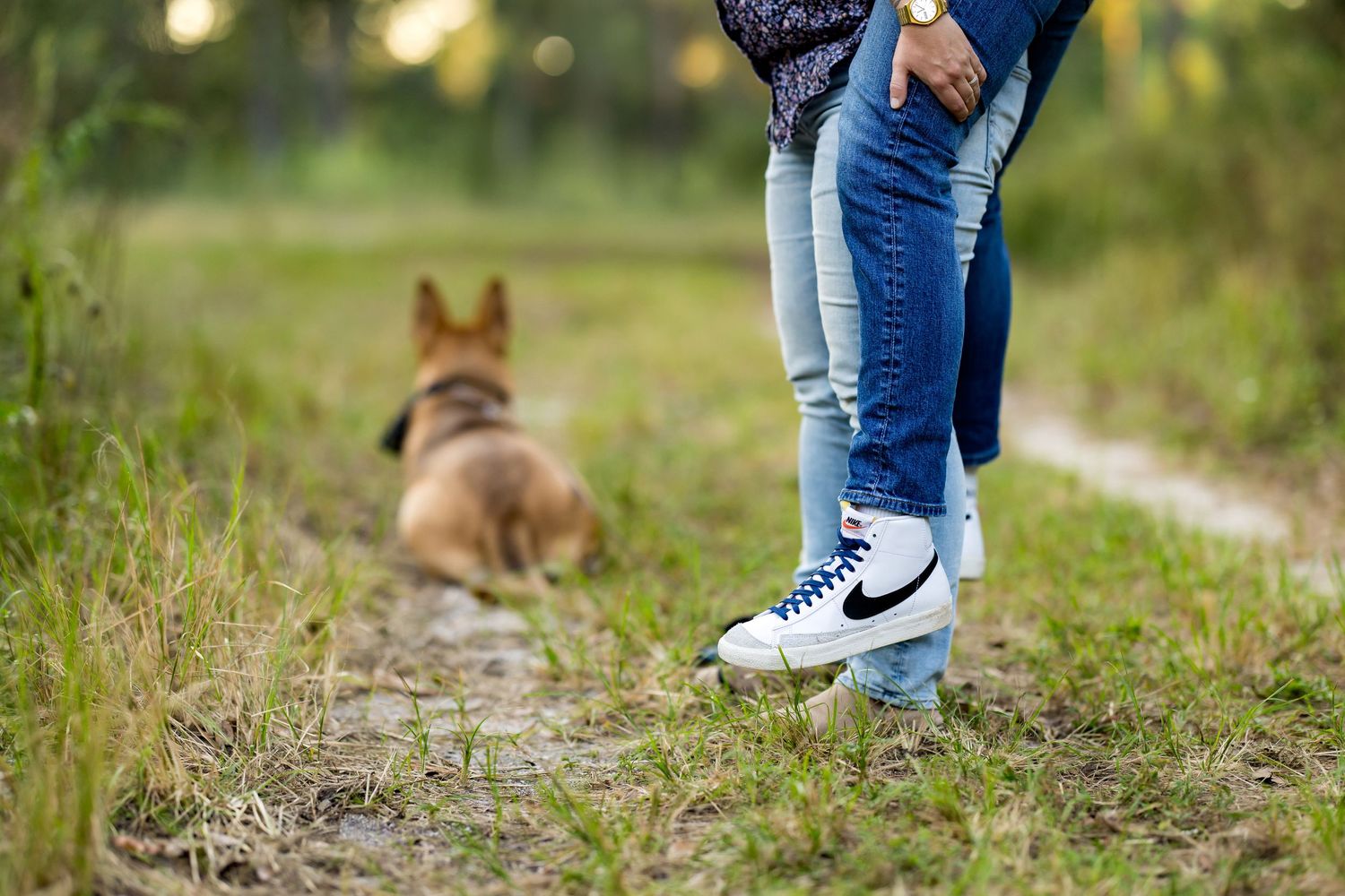 A small brown dog walks through a grassy path while someone in jeans and sneakers follows behind.