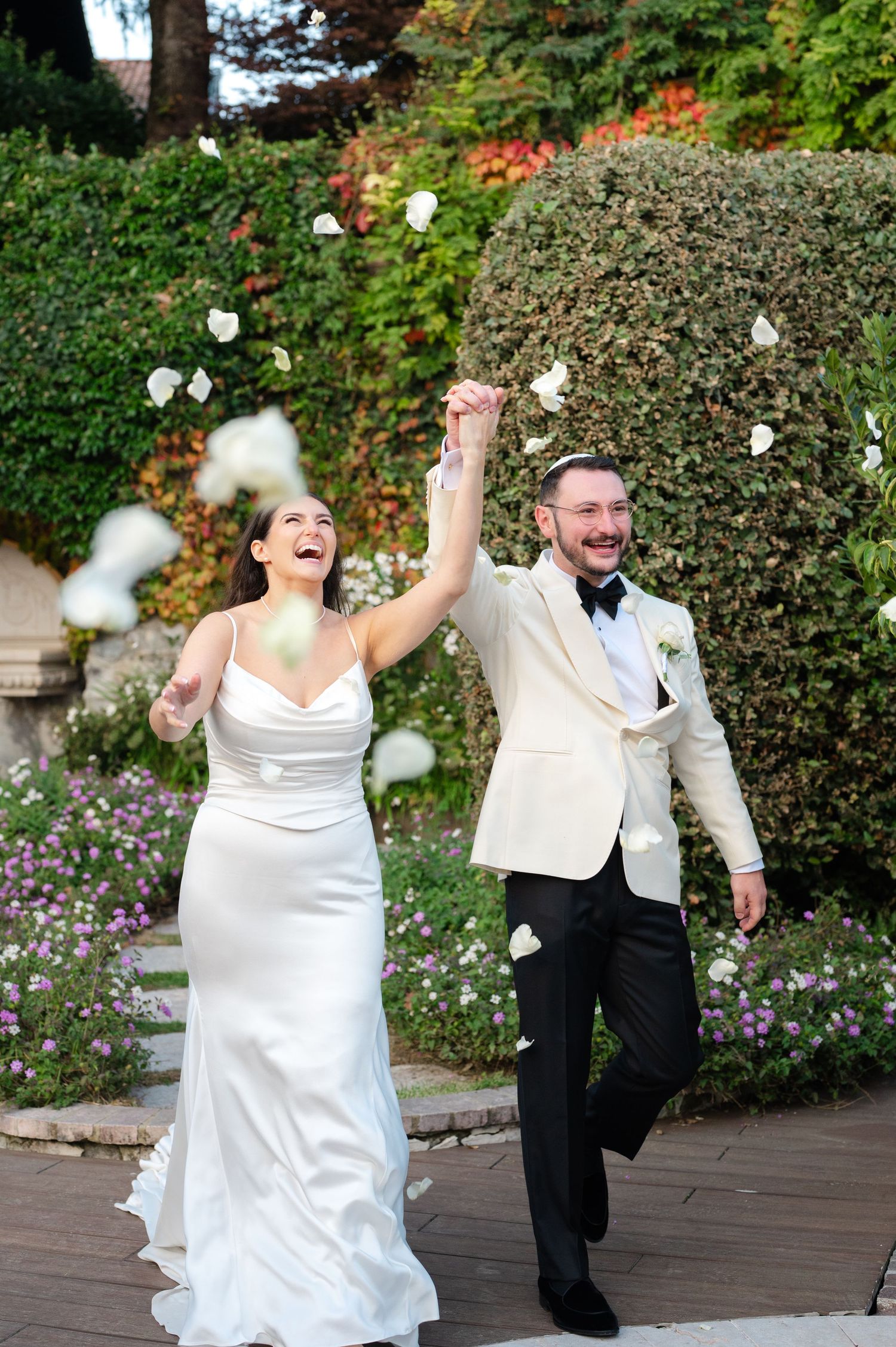 A playful wedding couple throws cotton balls while dancing and laughing together on a wooden deck surrounded by greenery.