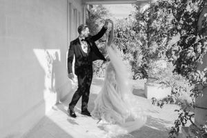 A dancing couple in wedding attire twirls in bright sunlight on a white veranda.