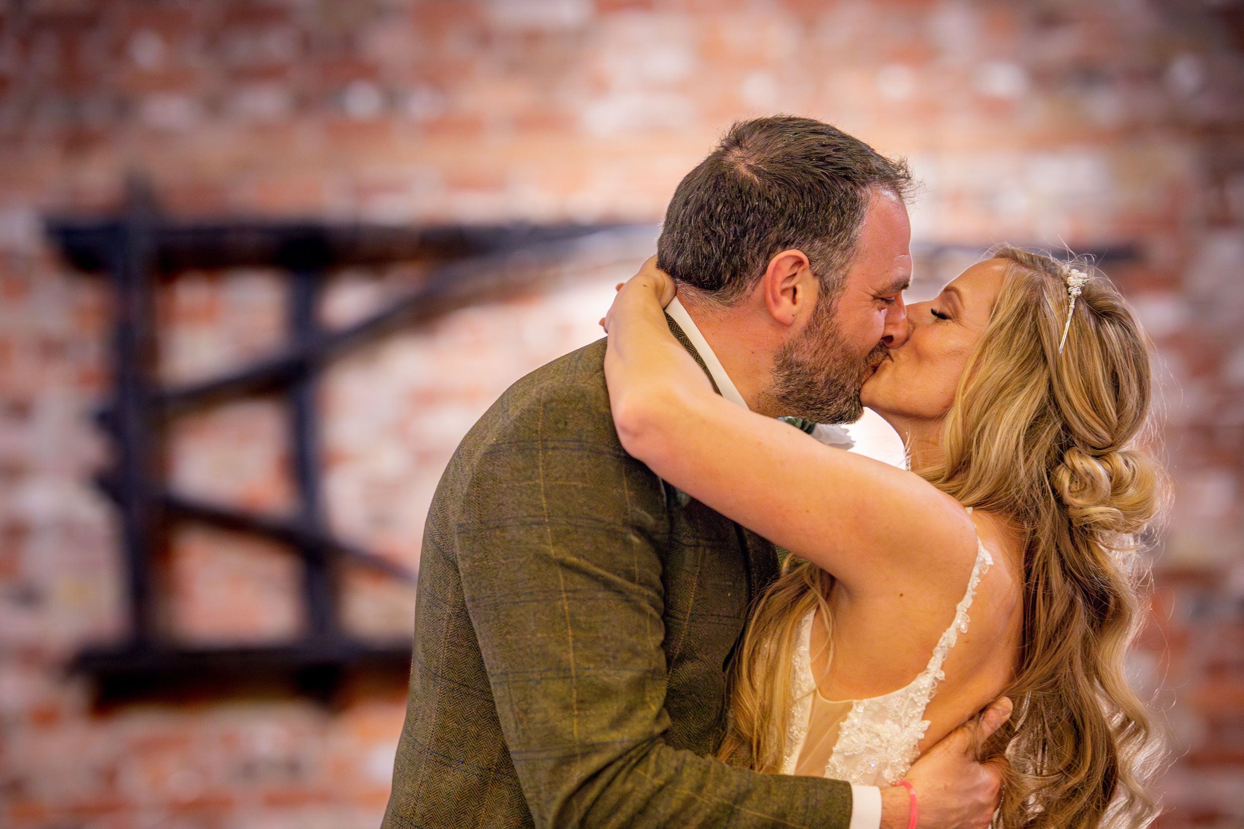 A romantic wedding kiss between a couple against a rustic brick wall backdrop.