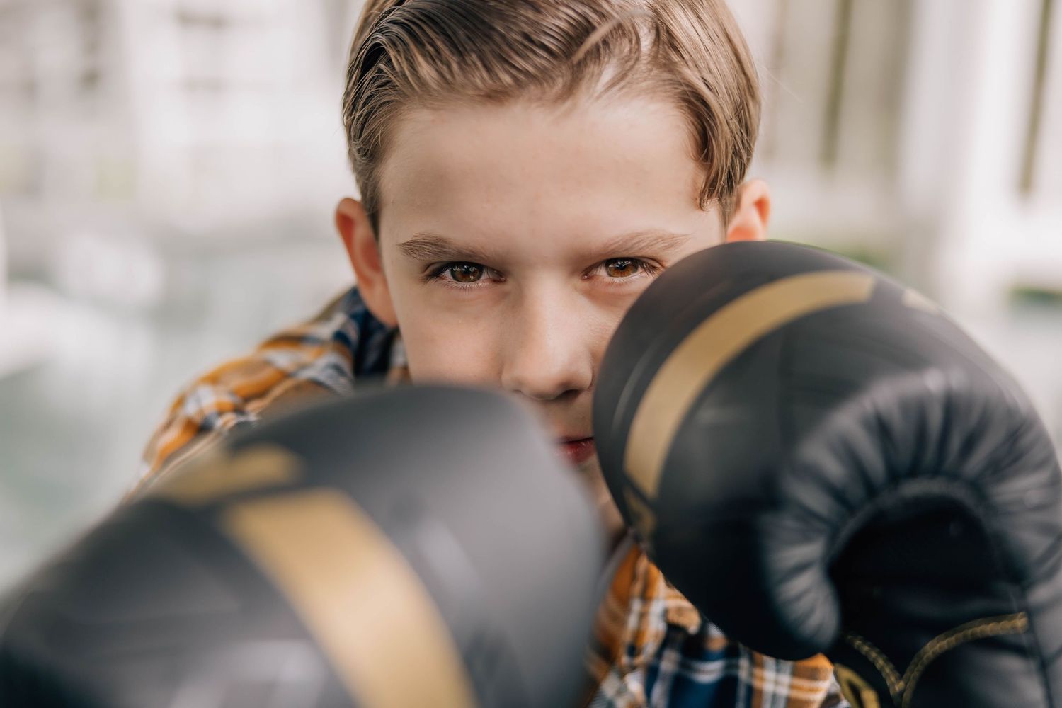 Close-up portrait of a young boy in a plaid shirt aggressively posing with black boxing gloves toward camera.