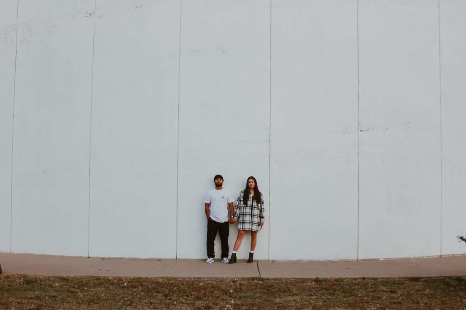 Couple standing apart in front of a large white wall outdoors.